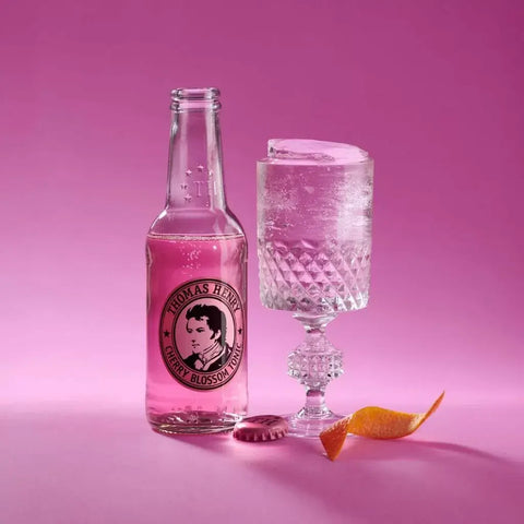 Thomas Henry Cherry Blossom Tonic 200ml bottle and glass with pink drink, garnished with citrus peel on a pink background.