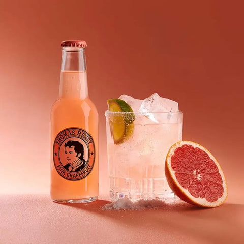 Thomas Henry Pink Grapefruit Tonic 200ml bottle with glass of ice and lime on a pink background, showcasing refreshing premium mixer.