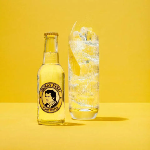 Thomas Henry Tonic Water 200ml bottle next to a glass with ice and lemon, set against a yellow background.