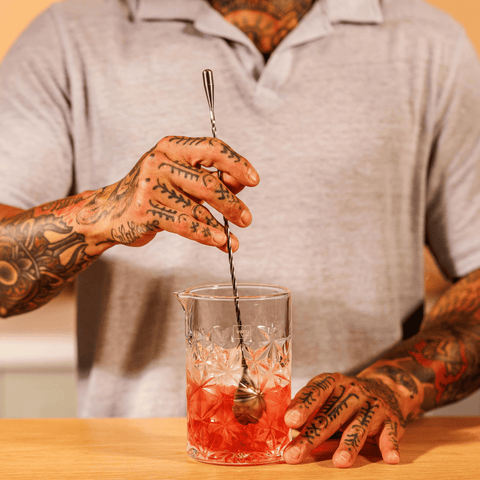 Bartender using Vacu Vin Bar Spoon Stainless Steel to mix a cocktail in a glass with tattooed hands.